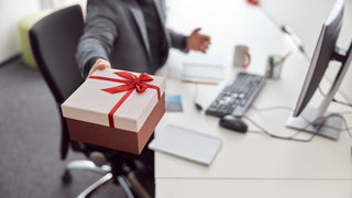 An office worker is sitting at their desk and is offering a gift wrapped box towards the camera.