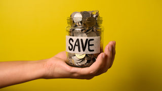 Clear glass jar with a label that says save, and is filled with coins.  An arm holds the jar up on a yellow background.