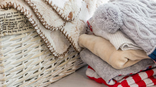 A white basket is filled with soft beige blankets, with a pile of baby clothes and a winter hat beside it.