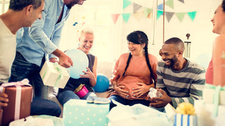 Woman with long brown hair in braids is surrounded by friends celebrating a baby shower.  She has her hands on her baby bump.