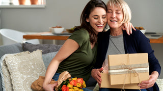 Young woman with dark hair and a green tshirt is holding tulips on her lap while sitting on a living room couch.  She is half hugging an older woman with short blonde hair holding a brown paper wrapped gift tied with a rustic bow and twine.