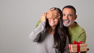 Husband hides his wife's eyes as he surprises her with a gift box wrapped in red ribbon