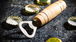 Bottle opener with wooden handle sits on black scratched surface with bottle caps surrounding it.