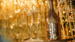 Golden background with 2 tall stemmed champagne glasses in the foreground, sitting beside a gold sequin covered champagne bottle.