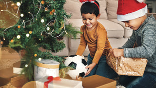 Kids sit beside their Christmas tree opening gifts.  One has just opened a soccer ball, and the other has just started opening a gold wrapped gift package.