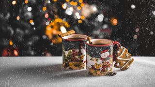 Two Christmas themed mugs sit side by side on a white table with a Christmas tree in the background, with snow falling, and a gingerbread tree cookie leaning up against the mug.  Mugs are imaged with snowmen.