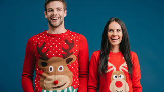 Man and woman standing on blue background wearing Christmas Sweaters with fun designs.