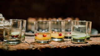 Assorted short clear glass drinking glasses sit on a wooden bar.  Some have remnants of orange juice and cranberries in them, some have gin and lime.