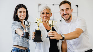 A young dark haired woman, a middle aged white haired woman and a young dark haired man all lean in and show  their various trophies.  2 are cups, one silver, and one gold, and one is a gold star.