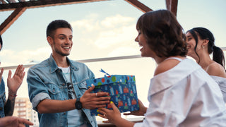 A young man is surprised at receiving a present in a blue box from his girlfriend in a beautiful outdoor setting.