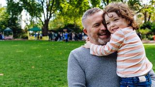 Father holds child with a park setting in the background.  Father has salt and pepper hair, child has brown wavy hair.