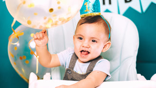 A young child celebrates their first birthday sitting in a white high chair with gold flaked balloons and a blue sparkly party hat.