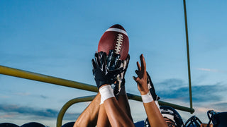 Hands of football athletes reach up and hold a football in the air at dusk outside.