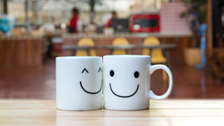 Two white ceramic coffee mugs have smily faces printed on them.  They sit in a coffee shop with the mugs in the foreground, and the shop blurred in the background.