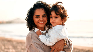 Woman with dark curly hair holding small child with dark hair on a beach.