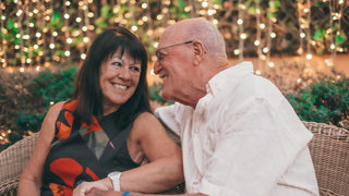 Older couple sitting on a rattan seat looking at each other happily with string lights in the background.