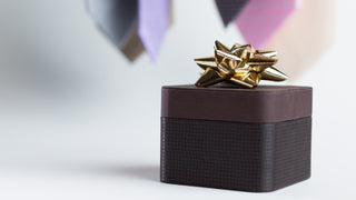 With the bottom of ties out of focus in the background, the foreground shows a brown leather style of gift box, with a gold bow on top.