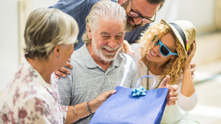 Elderly parents are gifted something in a paper bag with a bow by their older children outside in the sun.