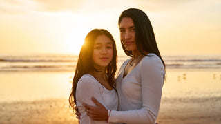 Two women, one younger and one older, stand embracing each other on a beach with a sunset in the background.