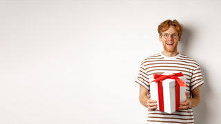 A red headed teenager with glasses and a white striped shirt, poses against a white wall holding a white gift box wrapped with a red bow.