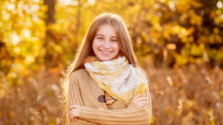 A teenage girl crosses her arms and smiles with a scarf and sweater in a golden forest light