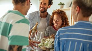 A group of friends are celebrating holding champagne glasses, a bottle, and white flowers