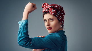 Woman wearing a red bandana with white polka dots wearing a jean shirt and is flexing for the camera.