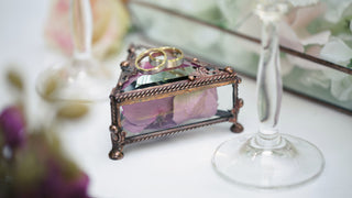 A small metal and glass jewelry box is sitting on a table with a flower in it, with wedding rings on top.  It is surrounded by some glassware.