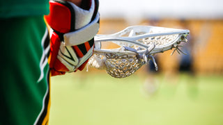 A close up of someone wearing a green uniform and red and white glove holding a lacrosse stick.