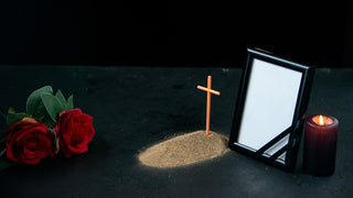 A picture frame is beside a candle and a cross with a red rose, beside a small gravesite.