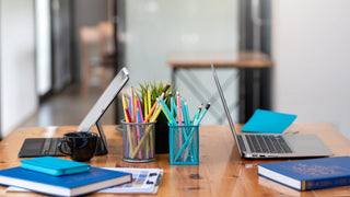 A plain wooden desk has pencil holders and laptops on it, with books in the foreground.