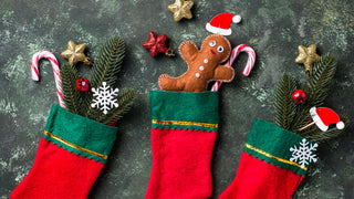 3 Red and Green felt Christmas stockings sit on a chalk background with various Christmas greenery, candy canes and felt gingerbread men sticking out the tops of each stocking.