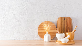 A light wooden table with a white wall.  Sitting on the table are a circular and rectangular cutting boards, and some mortar and pestles.