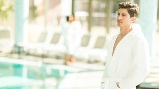 Young man stands looking in the distance with a pool in the background.  He is wearing a white bath robe.