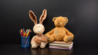 A brown stuffed bunny and stuffed teddy bear are arranged on a black background with a pencil holder beside the bunny and the bear sitting on top of some stacked notebooks