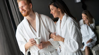 A couple stands holding coffee mugs with their arms linked, while looking out the window at a spa.  They are wearing plush white bath robes.