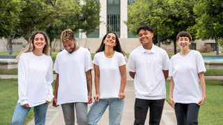 A group of young adults wear white tshirts in front of an academic looking building covered by green trees.