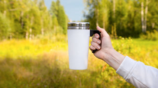 Hand wearing a white sweater holds a white travel coffee mug with a black handle on a field background.