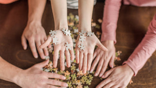 Hand are holding puzzle pieces above hands that are completing a puzzle on the table below.