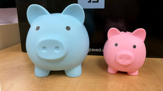 Two plastic personalized piggy banks sit in front of a black things engraved box.  One is larger and blue, the other is smaller and pink.