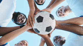 Group of male soccer players holding a black and white soccer ball while the camera takes a photo upwards of the team huddle.