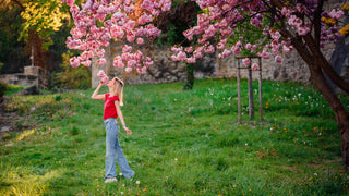 Blonde woman wearing a red tshirt and jeans reaches up to smell the flowers on a cherry blossom tree in the spring.