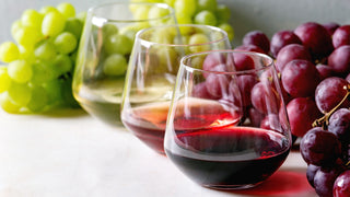 3 Stemless Wine Glasses sit on a table surrounded by grapes, and are filled with red, rose and white wines.