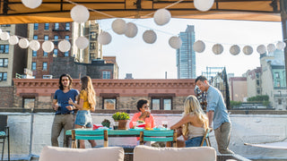 A group of friends getting together on a modern rooftop patio with garlands and teal furniture.  Drinks are in glasses on the patio table.