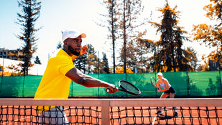 man in white hat and yellow shirt is playing tennis on a clay court. In the background is a female tennis partner.