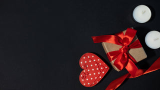 A small brown gift box is wapped with a silky red ribbon, and sits beside a red heart with polka dots and 2 tea candles.