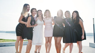 Group of women in fancy cocktail dresses pose outside for a photo wearing bachelorette party sashes.