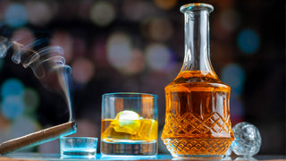 A cigar sits beside a standard straight edged whiskey glass with rocks, and a glass decanter.